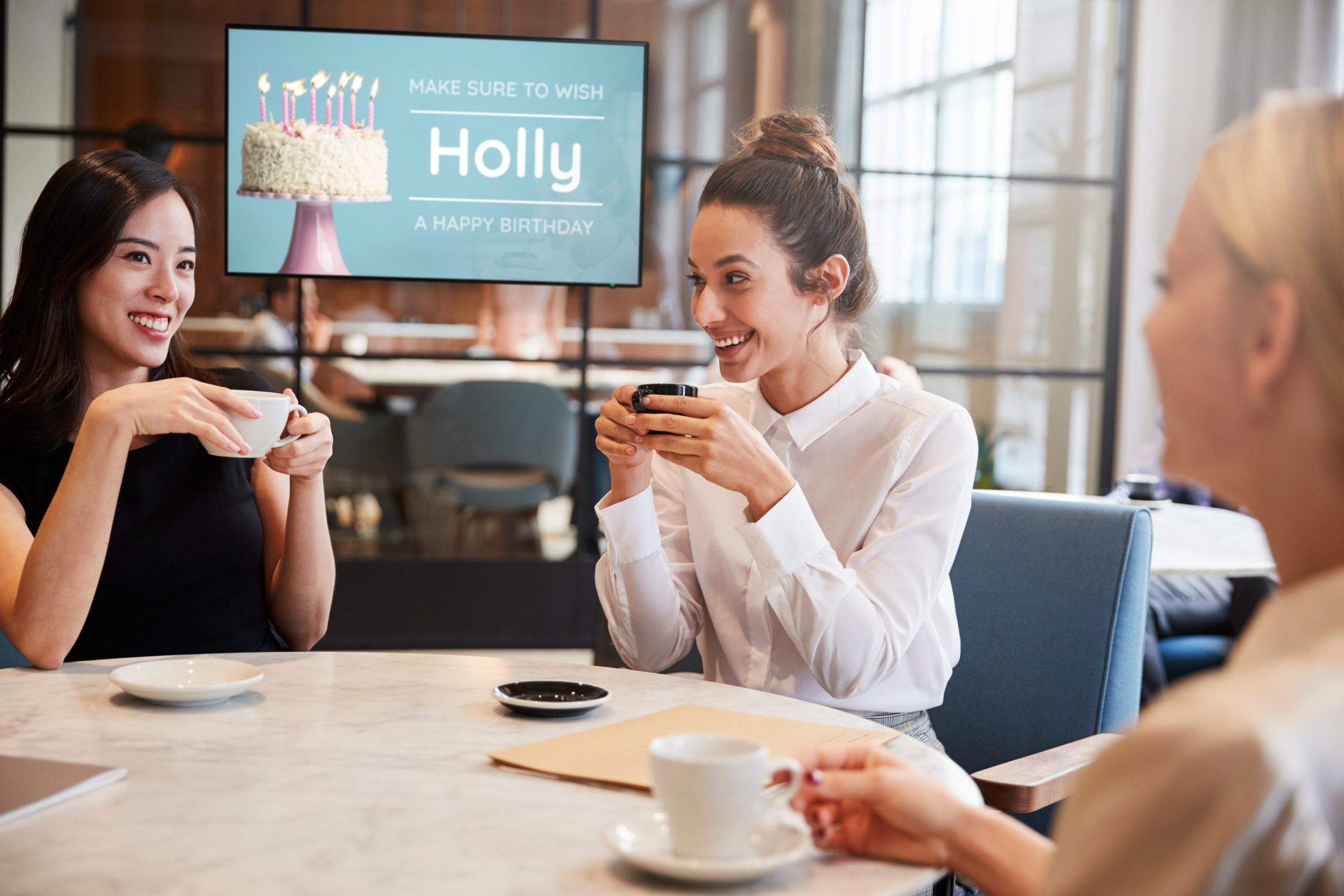 Three female coworkers sit around a table having coffee, a digital sign is in the background that reads "don't forget to wish Holly a happy birthday".