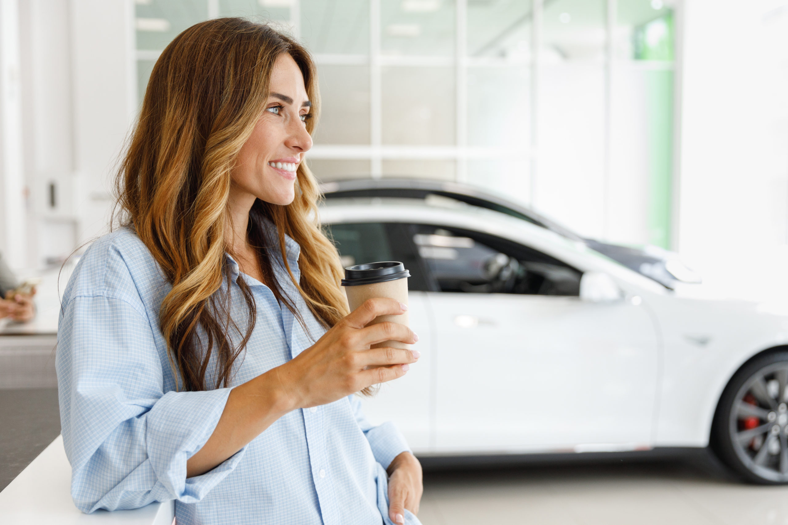 Minded woman customer female buyer client in shirt hold takeaway delivery craft paper cup coffee choose auto want buy new automobile in car showroom vehicle salon dealership store motor show indoor