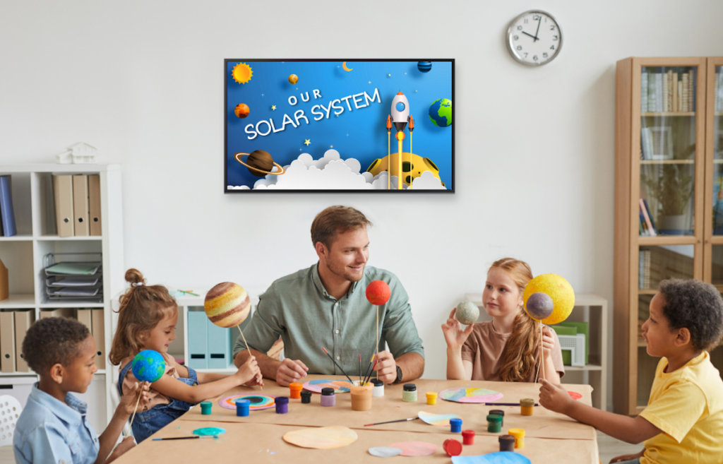 classroom with teacher and kids at table, a digital display hangs on the wall