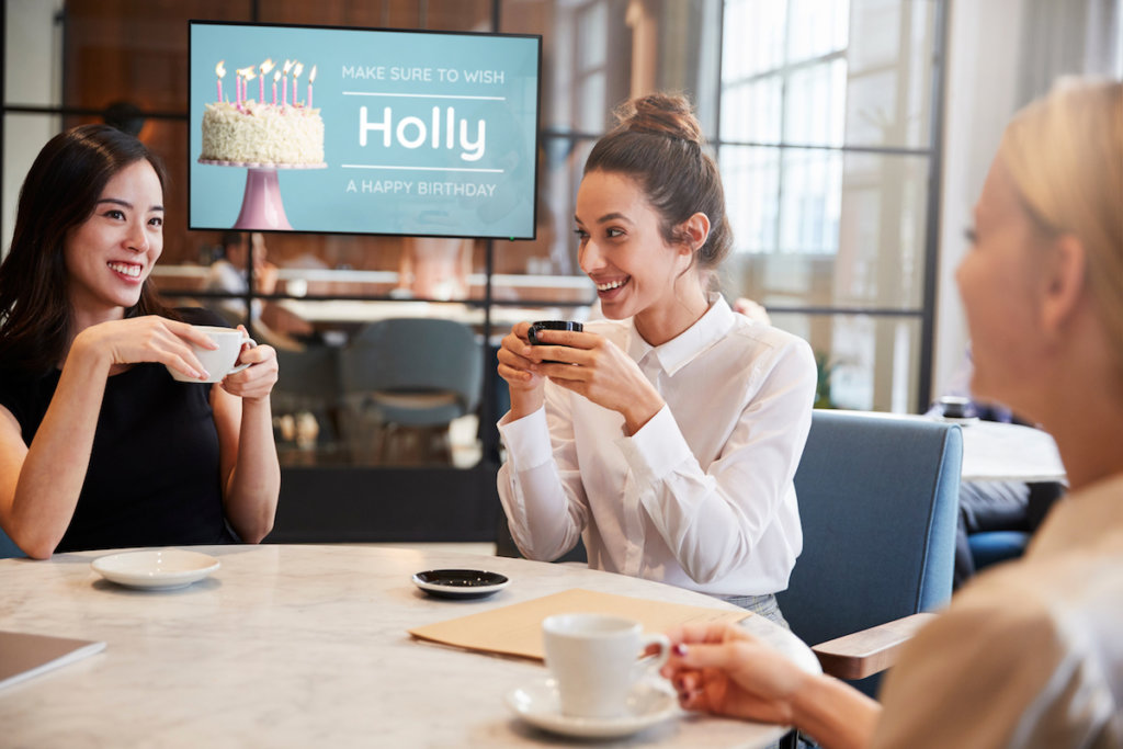 three workers sip warm drinks at a table break room with a display screen in the background