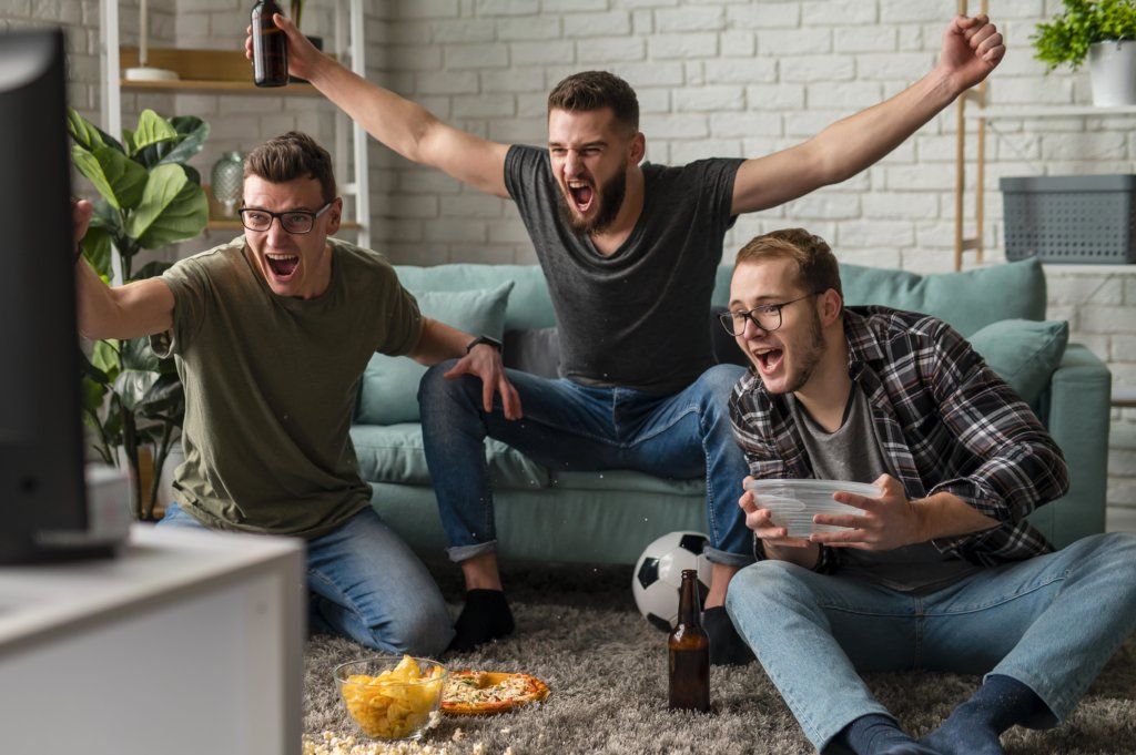 view from back of tv of the friends cheering towards the tv with snacks and a soccer ball on the floor