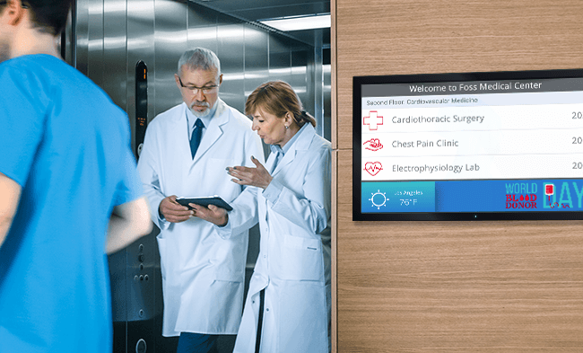 Doctors exit elevator next to digital directory.