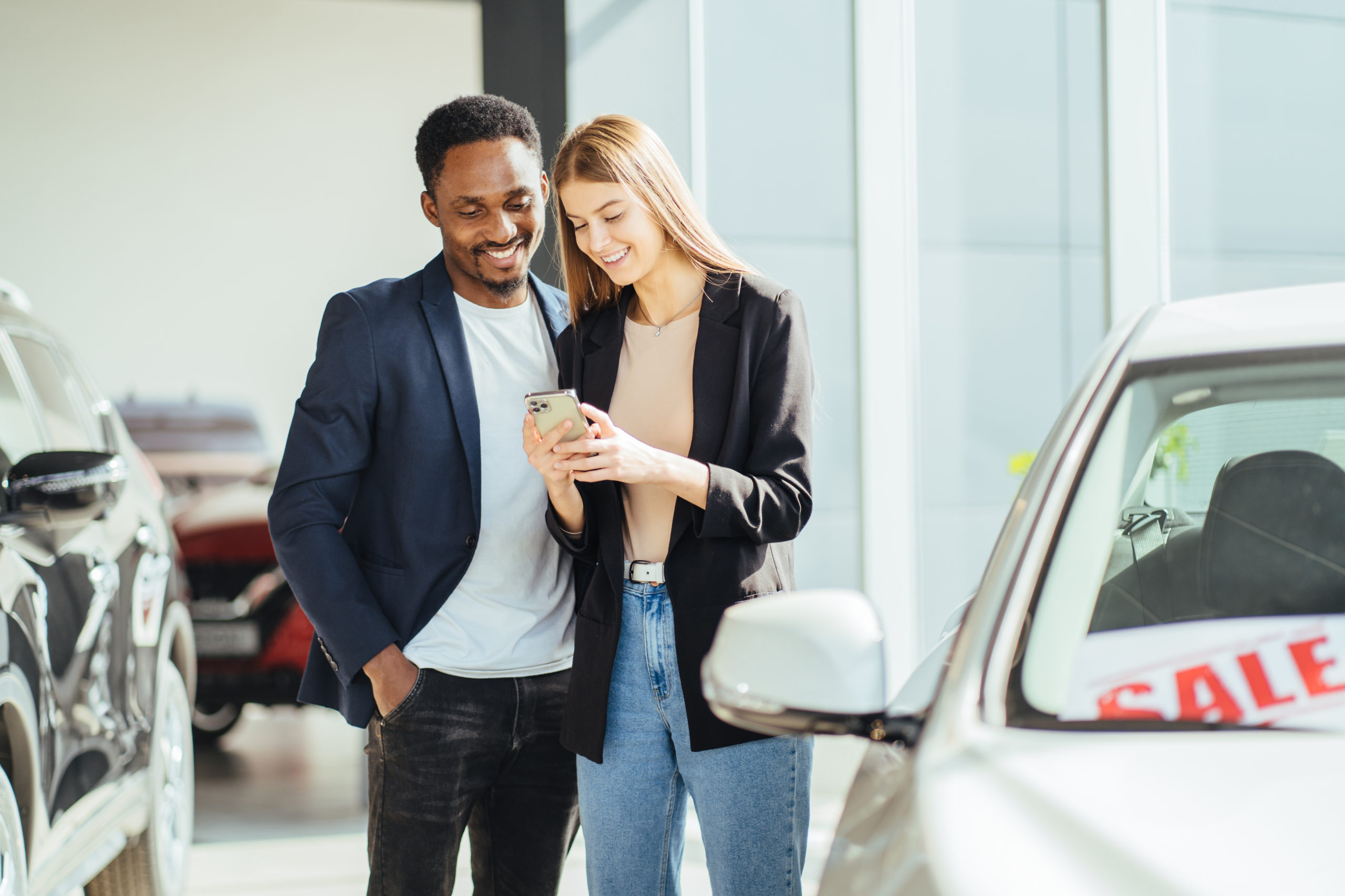 eautiful multiracial couple standing together at auto salon and using modern smartphone. African man and caucasian woman choosing new vehicle at showroom.